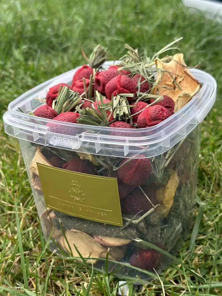 A clear plastic trap loaded with dried fruit, herbs and citrus peel, standing in grass at a stable yard
