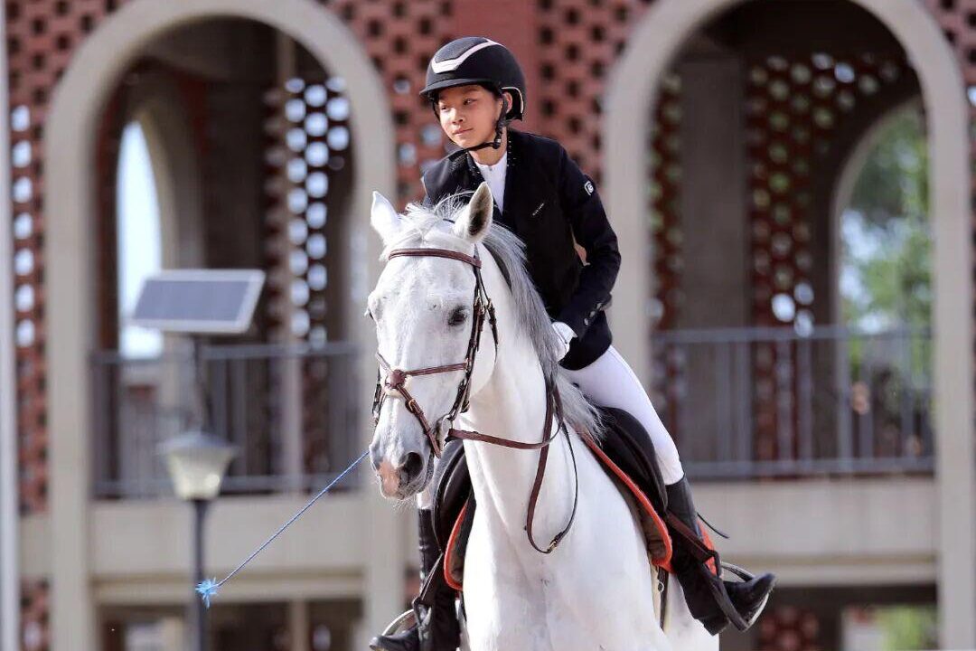 A young rider in a black helmet and jacket jumping a grey horse over a fence in the school arena