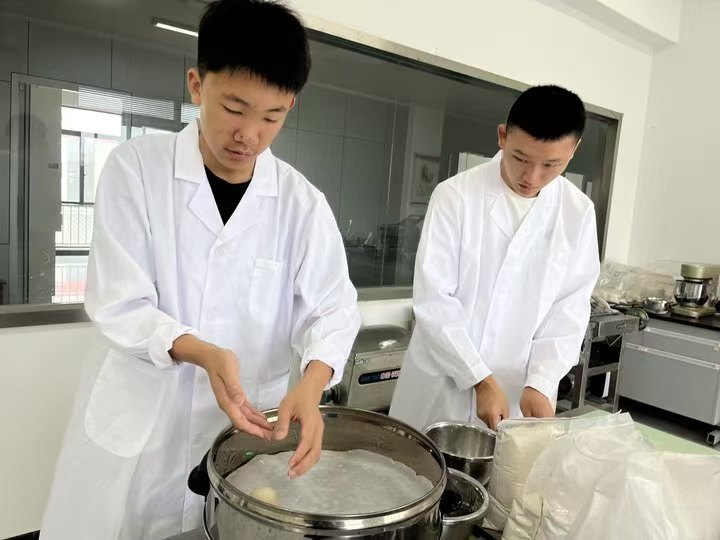 Two students in white lab coats preparing food beside a steamer in the school kitchen laboratory
