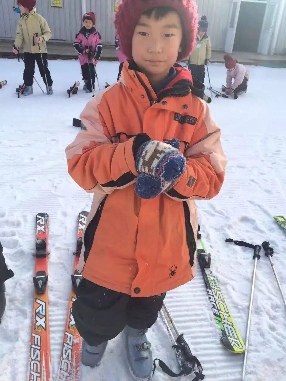A young boy in a bright orange ski jacket standing on snow with skis beside him