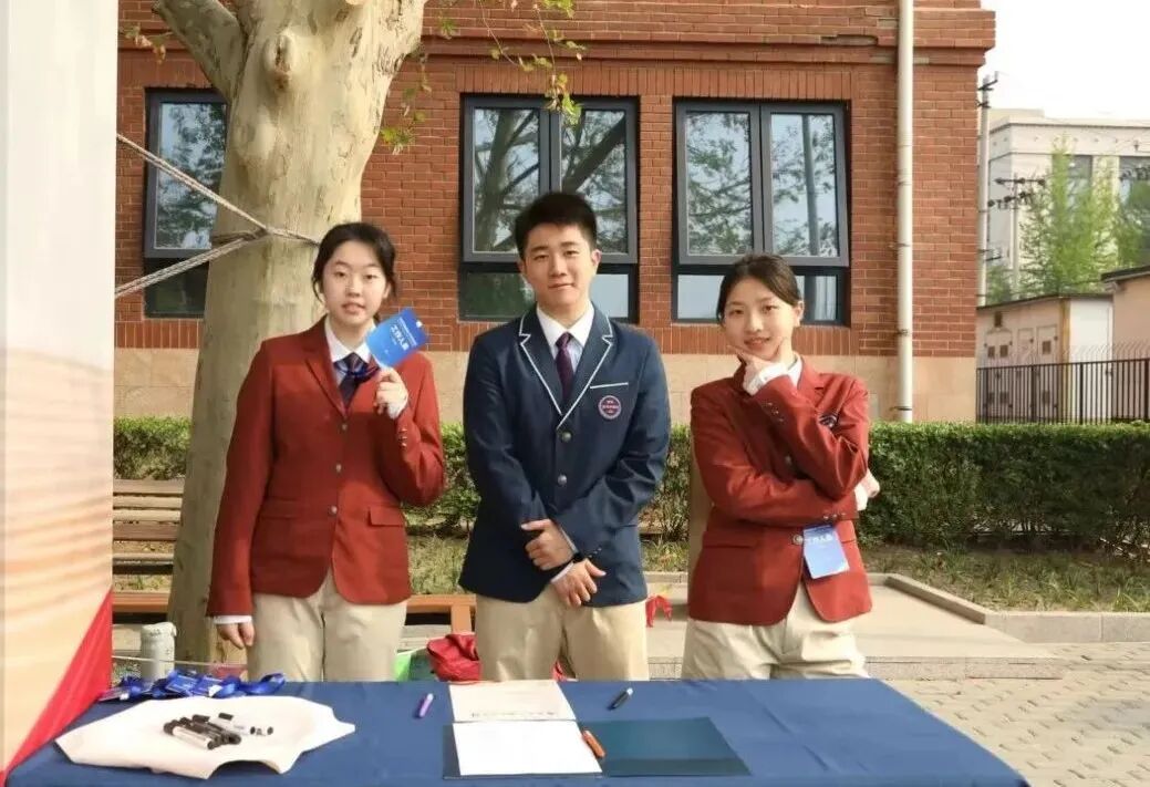 Three students in red blazers standing at an outdoor information booth on campus
