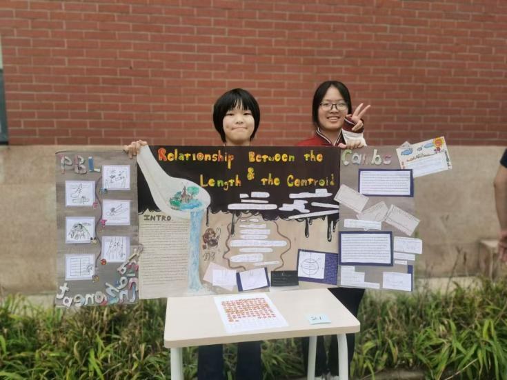Two students holding up their PBL poster on the relationship between length and control, set up on a table in front of a brick wall