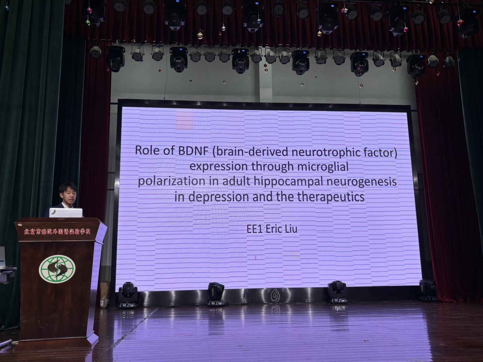 Eric Liu at the lecture podium presenting his PBL research, with his slide on the role of BDNF expression through microglial polarization in adult hippocampal neurogenesis in depression and the therapeutics on the auditorium screen