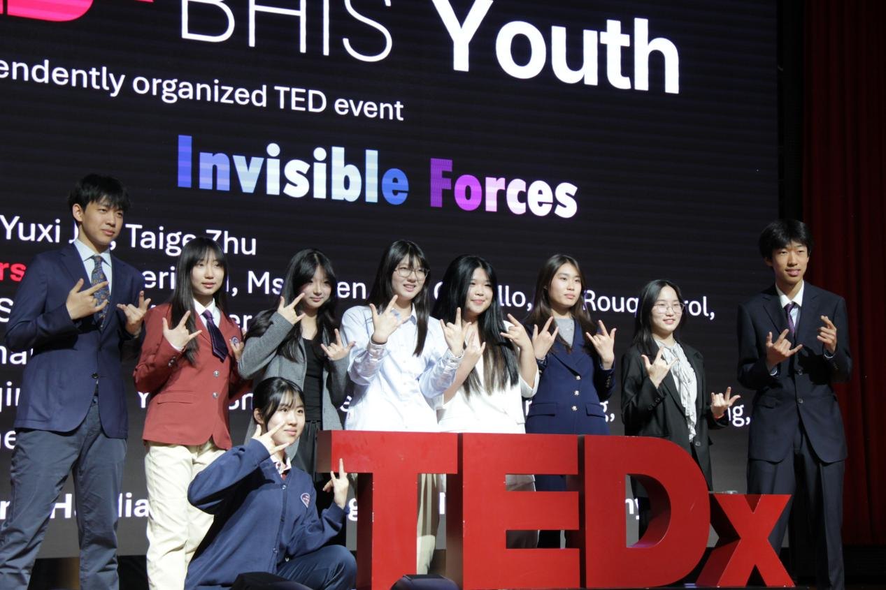 The 2025 TEDx BHIS team posing with the TEDx sign in front of the Invisible Forces stage screen