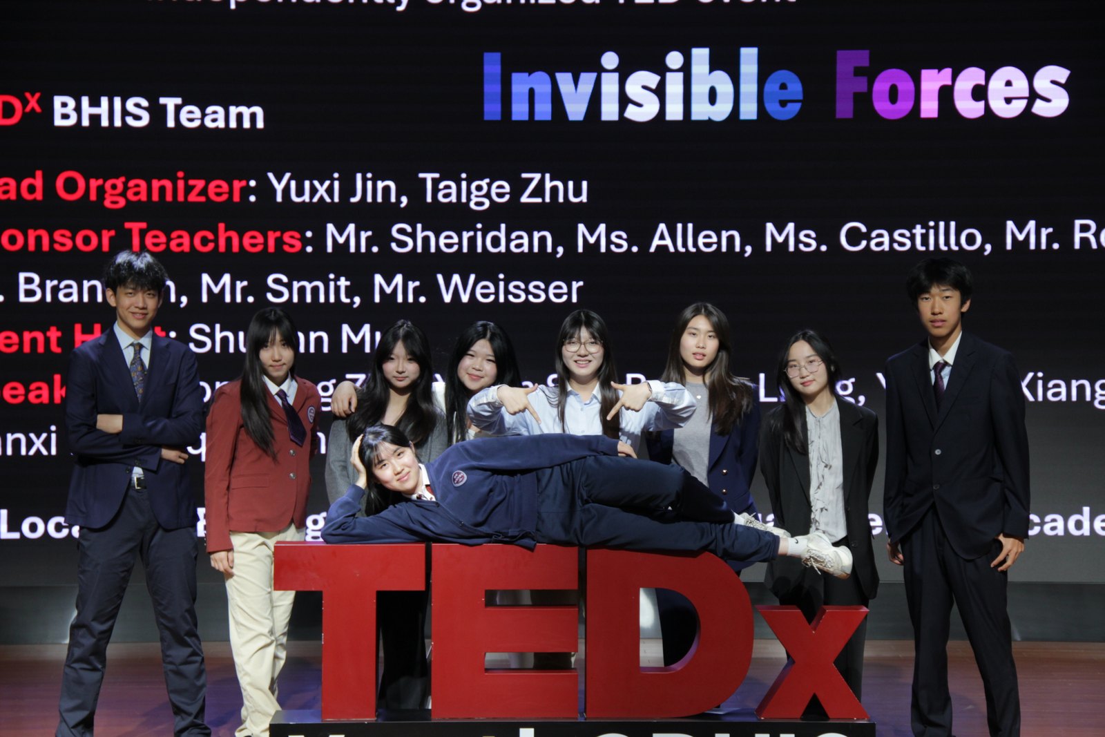 The 2026 TEDx BHIS team posing with the TEDx sign in front of the Invisible Forces stage screen