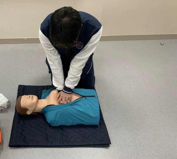 A student kneeling over a first-aid training manikin, arms straight and hands stacked on the centre of the chest