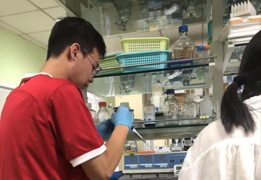 A student in a red shirt and blue nitrile gloves loading samples with a pipette at a crowded laboratory bench