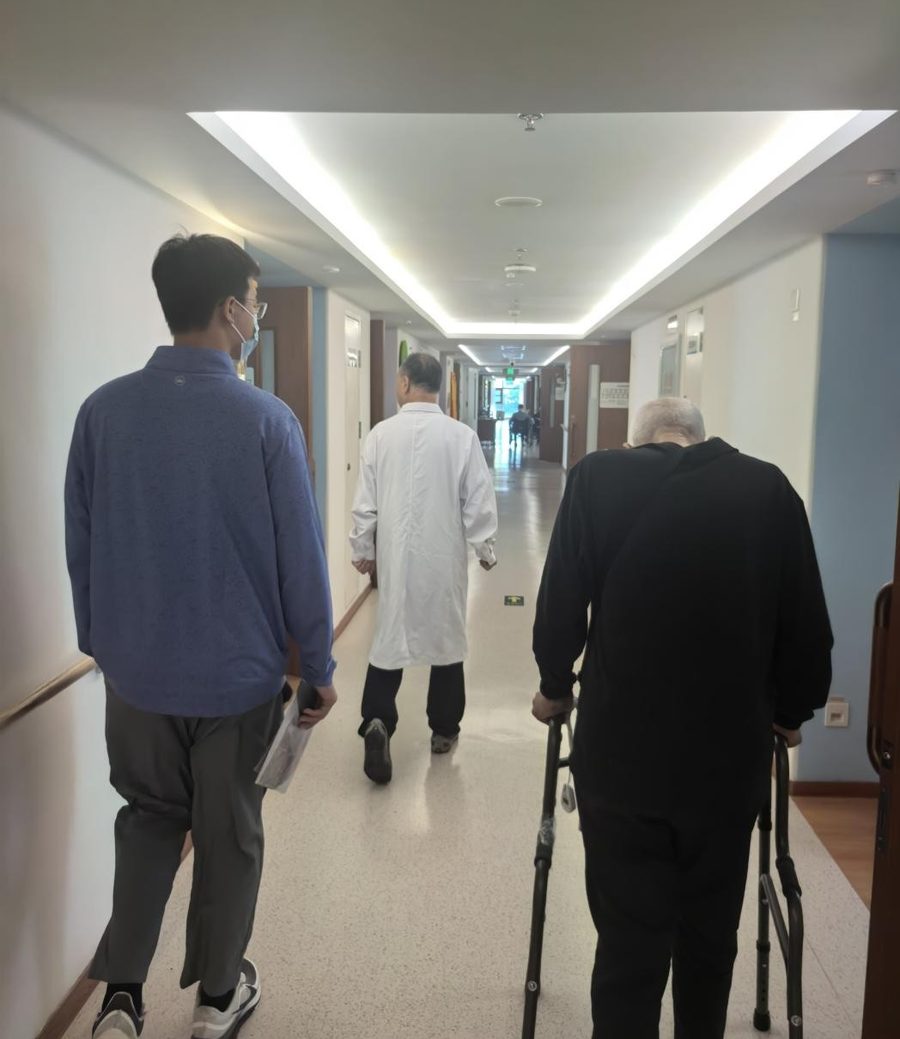 A student walking beside an elderly resident with a walking frame down a brightly lit care-home corridor